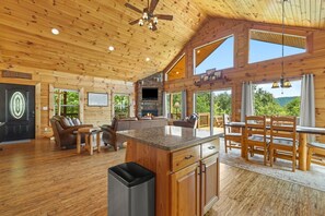 A cabin interior with a stone fireplace, wooden furniture, and large windows offering a view of the outdoors. - Honeymoon Ridge- Luxury Mountaintop Cabin (Bryson City)
