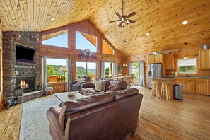 A spacious living area with a fireplace, leather sofa, and a large window overlooking a mountainous landscape. - Honeymoon Ridge- Luxury Mountaintop Cabin (Bryson City)