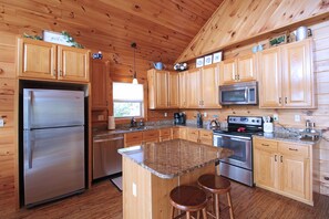 A wooden cabin kitchen with a stainless steel refrigerator, granite countertops, wooden cabinets, a microwave, a stove, and two bar stools. - Honeymoon Ridge- Luxury Mountaintop Cabin (Bryson City)
