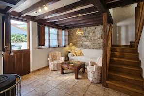 A cozy living room with a stone wall, wooden beams, a sofa, two patterned armchairs, a coffee table, and a staircase.