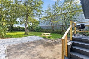 Eine Beton-Terrasse mit Picknicktisch, Grill und einer Holztreppe zu einem Gebäude.