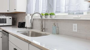 Eine Küche mit Spüle aus Edelstahl, weißen Schränken, einer Mikrowelle und einem Fenster mit weißen Vorhängen.