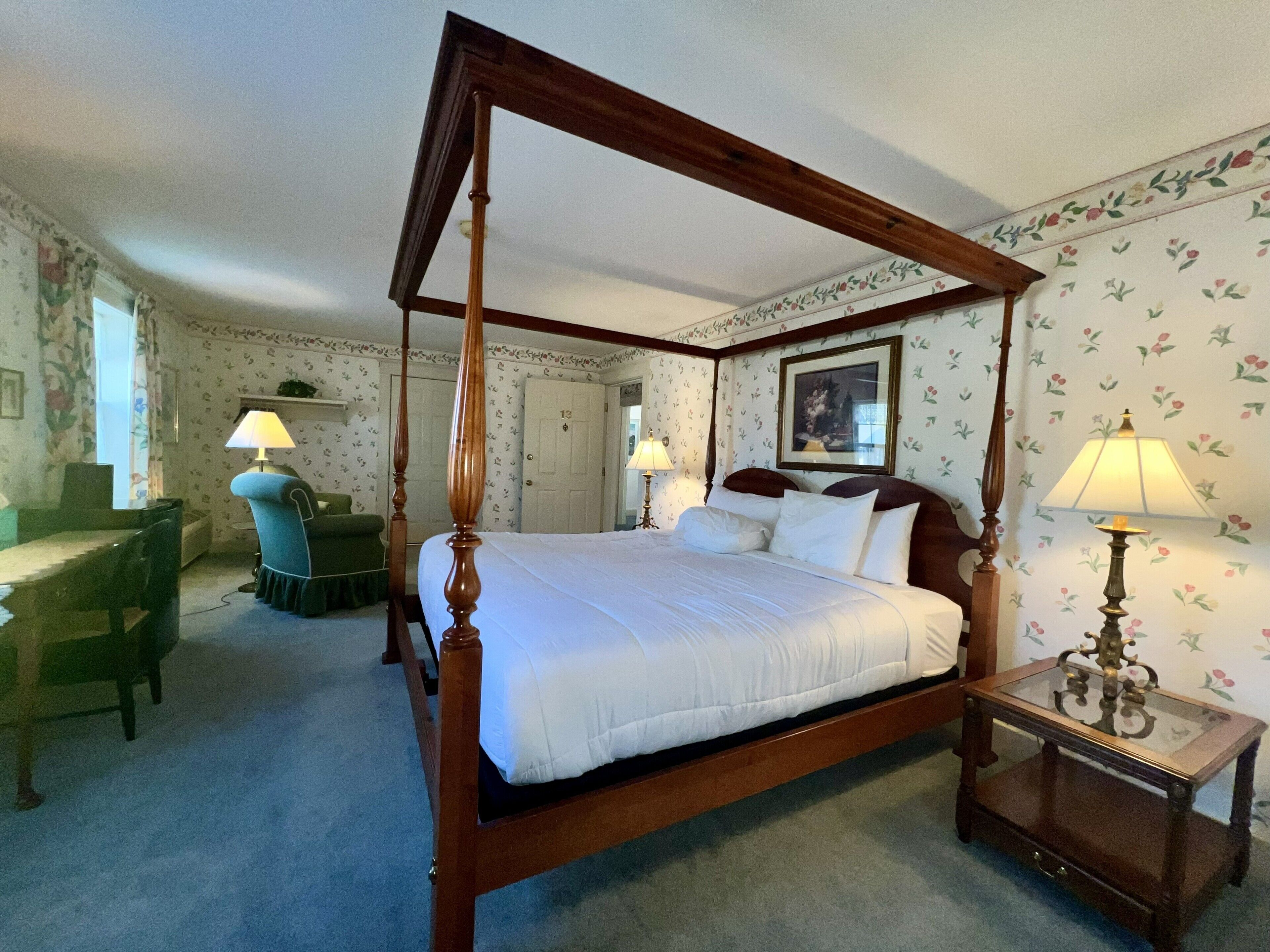 A four-poster bed with a wooden frame, a desk with a chair, two lamps, and floral wallpaper.