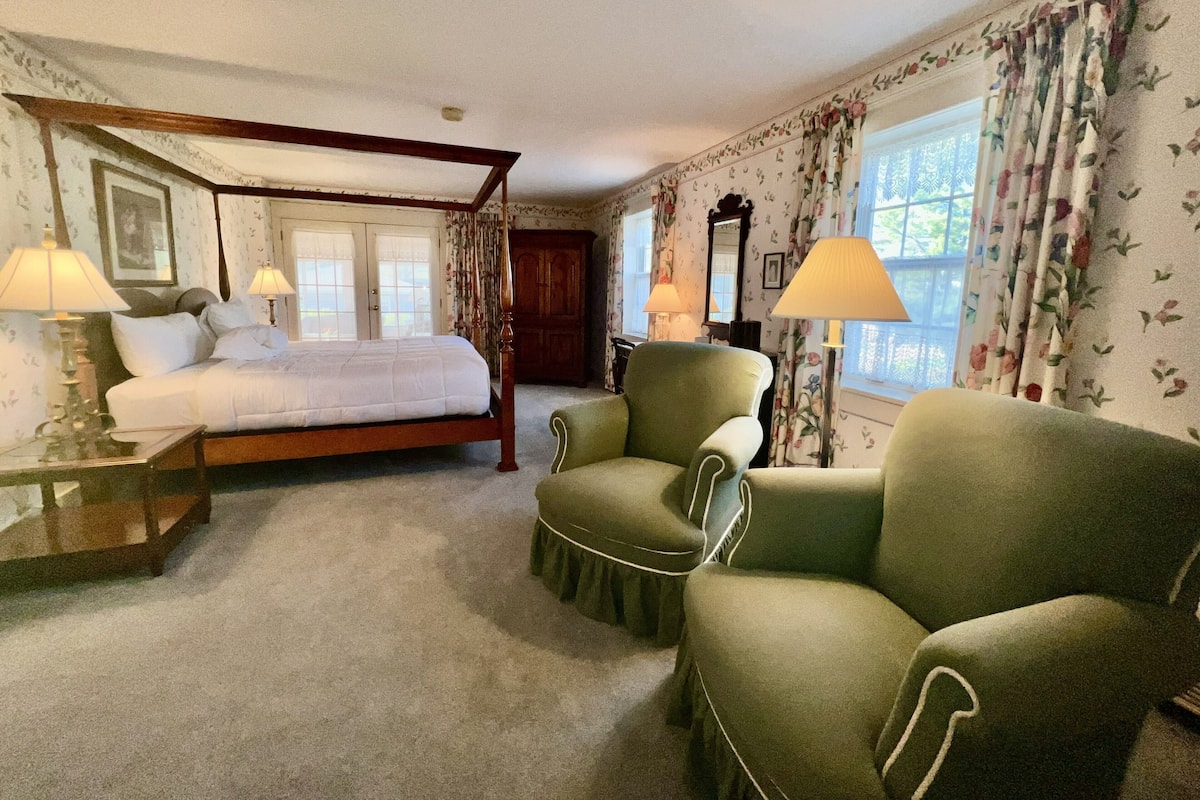 A bedroom with a four-poster bed, two armchairs, a wooden nightstand, a dresser, and a window with floral curtains.