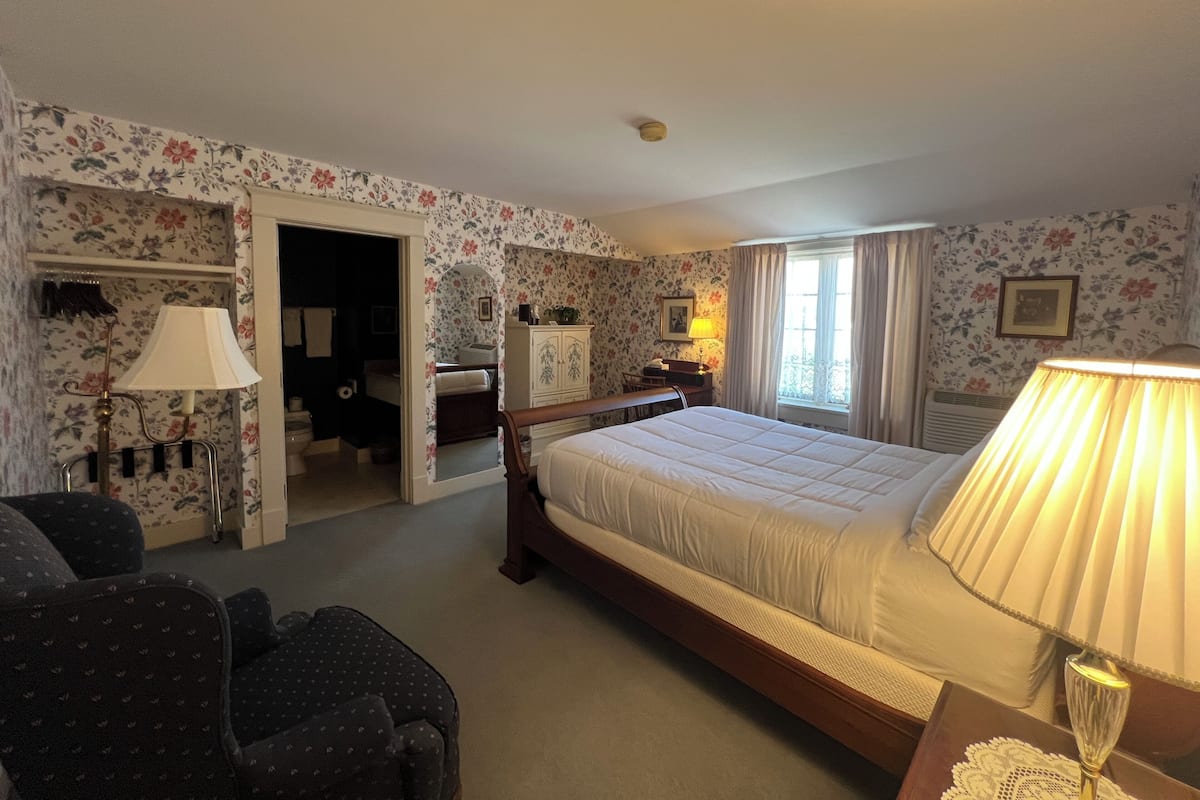 A hotel room with a bed, a desk, a chair, a lamp, and a door leading to a bathroom.