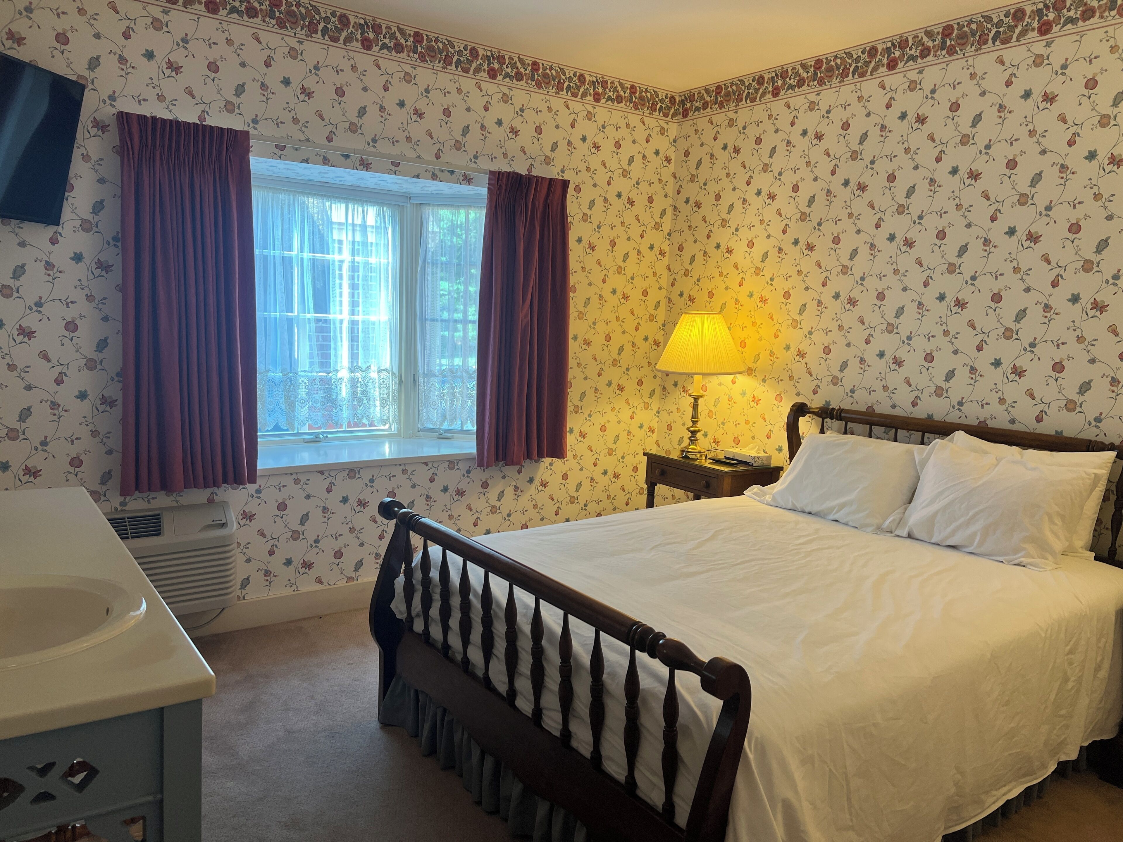 Een slaapkamer met een bed, een nachtkastje met lamp, een raam met gordijnen en een aan de muur bevestigde televisie.