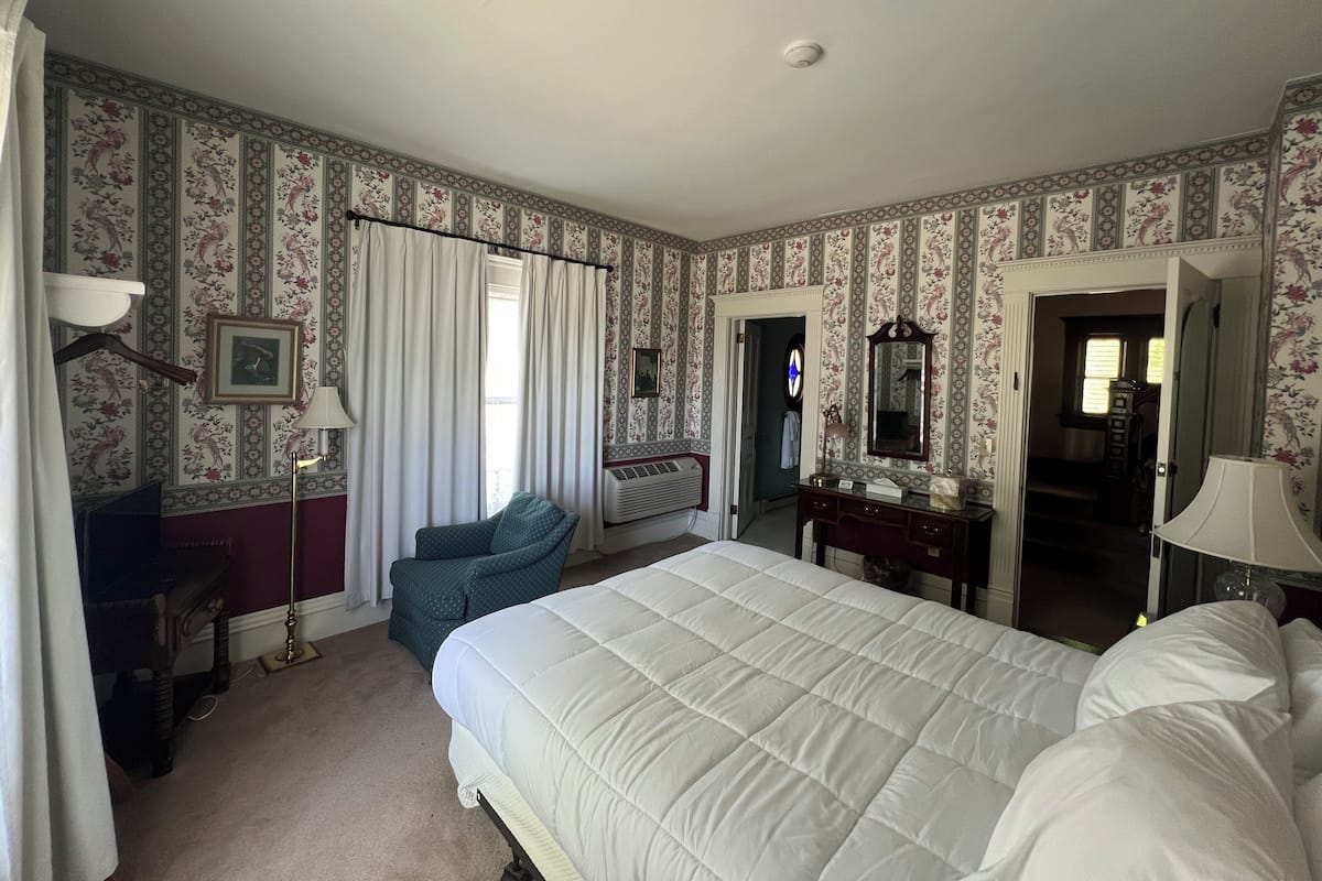 A bedroom with floral wallpaper, a bed, a chair, a desk, a mirror, and a radiator.