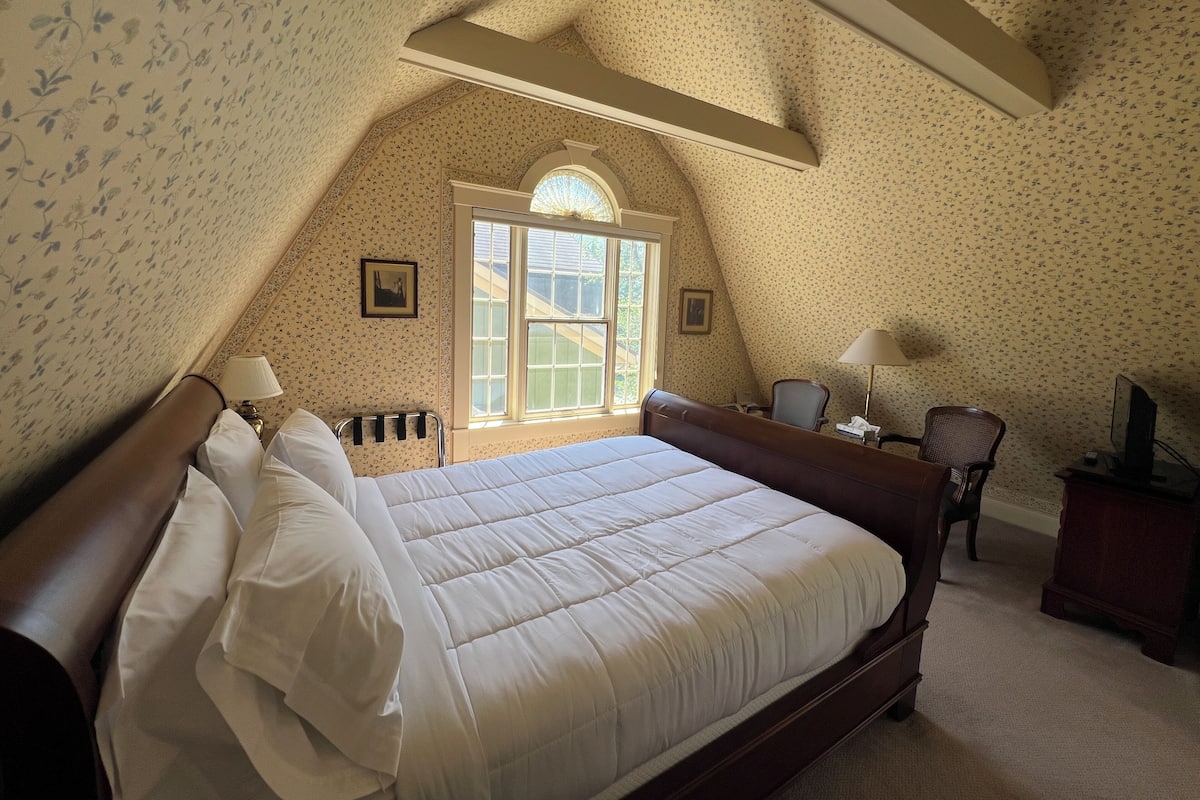 A bedroom with a large bed, a window, a chair, a desk, and a lamp.