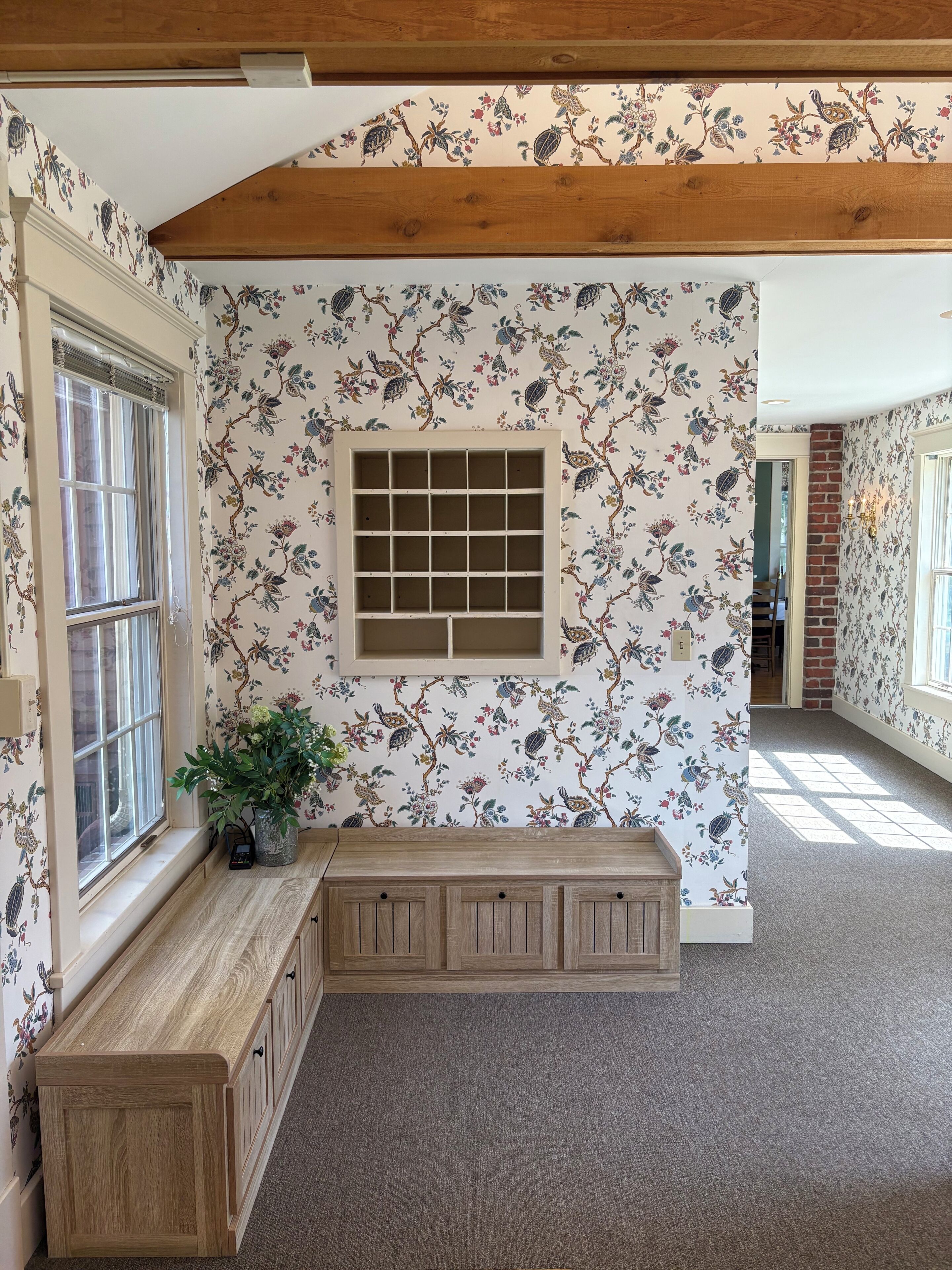 Une pièce avec un banc en bois, une fenêtre avec des stores et un papier peint à motifs floraux.