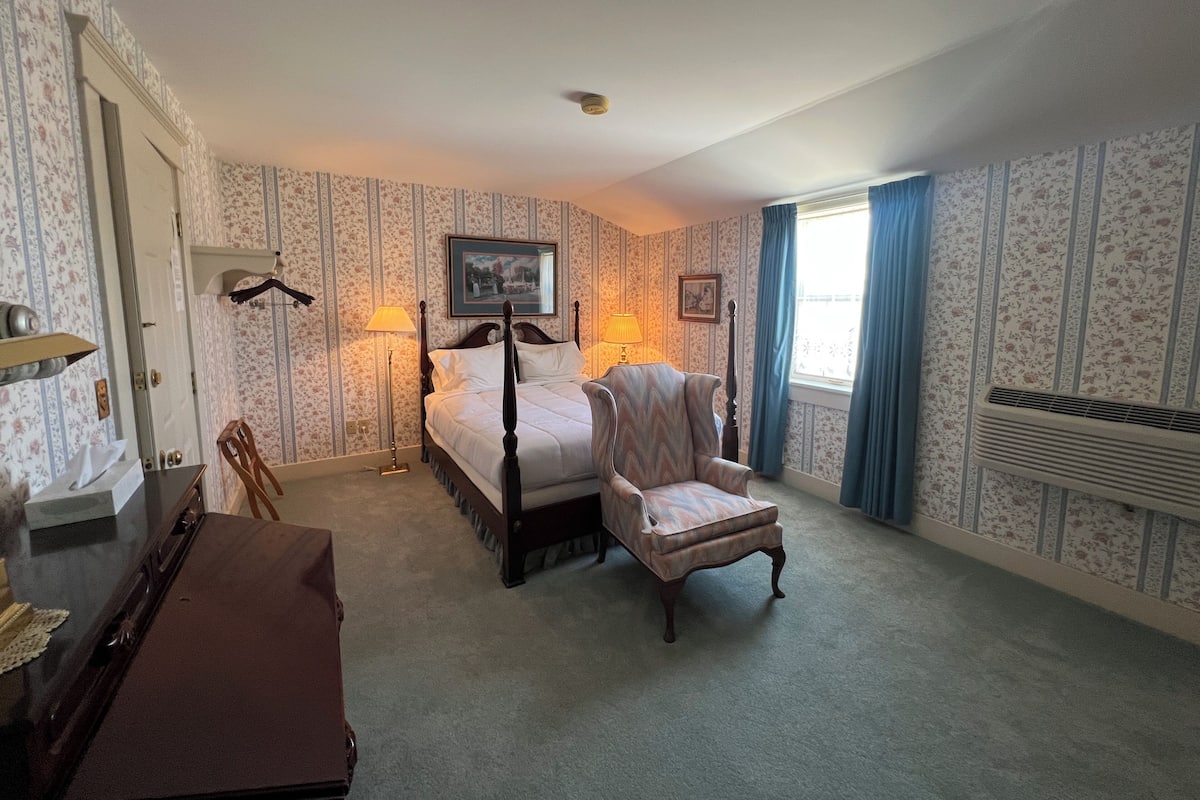 A hotel room with a bed, a chair, a desk, a lamp, a picture on the wall, and a window with curtains.