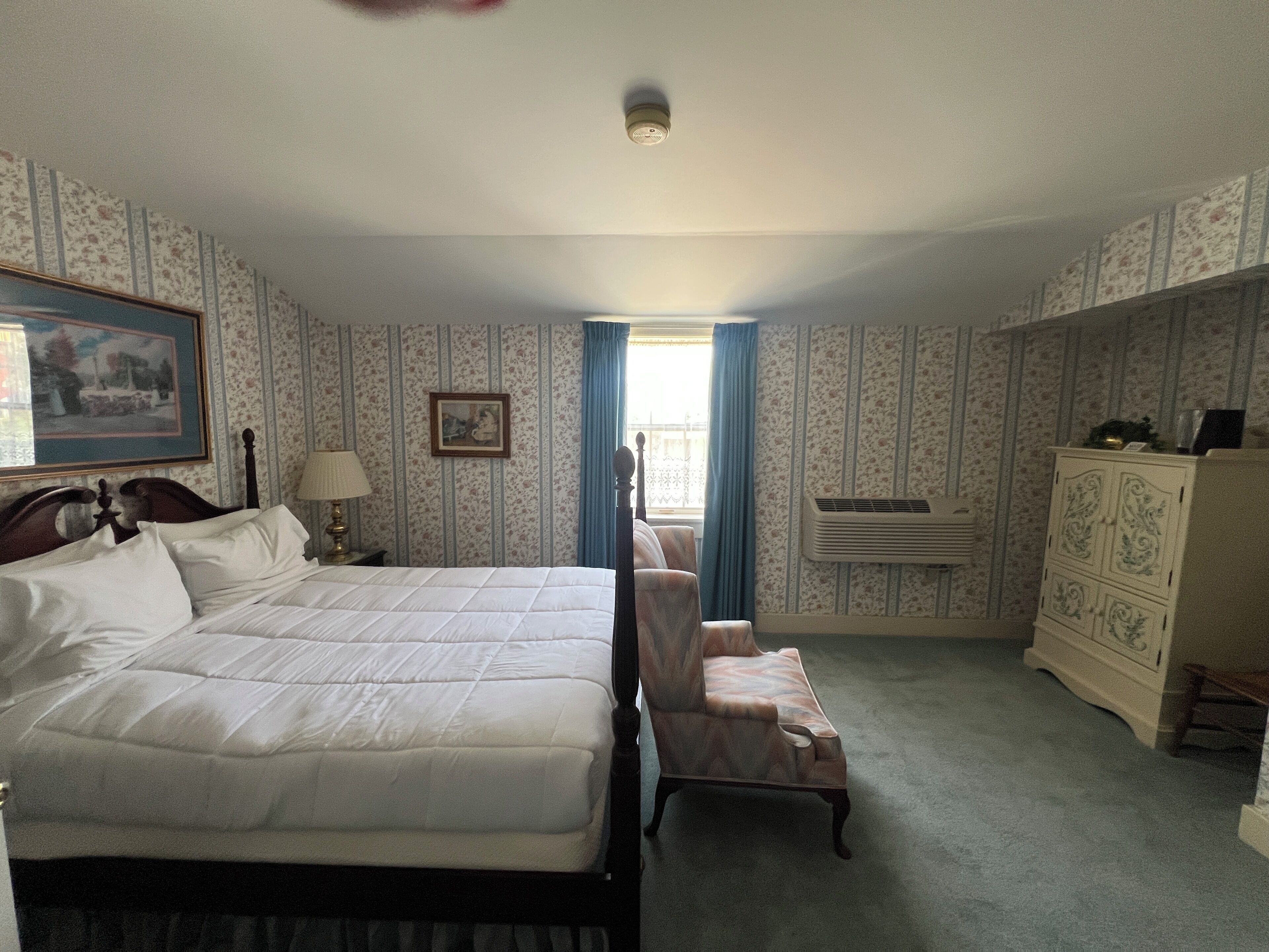 Habitación de hotel con una cama grande, una silla, un tocador, un cuadro en la pared y un equipo de aire acondicionado.