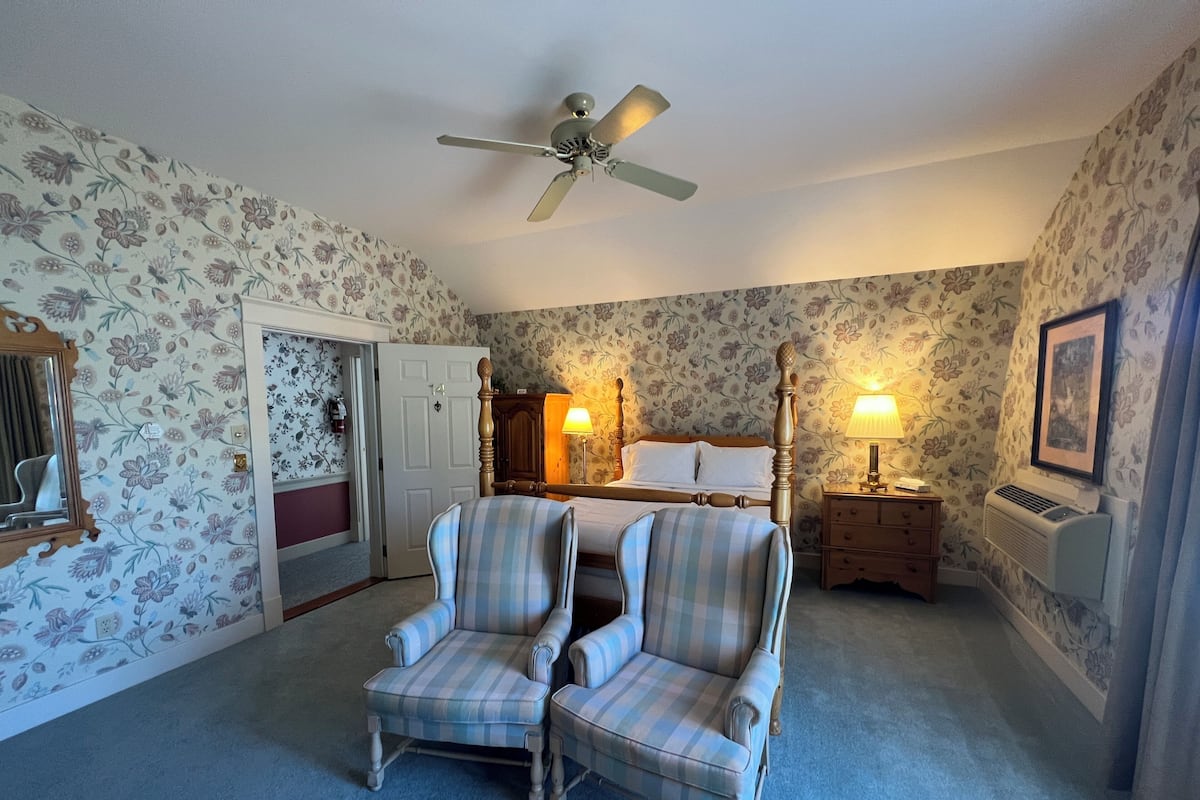 A bedroom with floral wallpaper, a bed with white linens, two armchairs, a ceiling fan, a nightstand with a lamp, and a radiator.