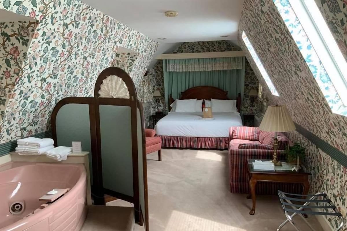 A hotel room with a large bed, a sitting area, a bathtub, and a window with a view.