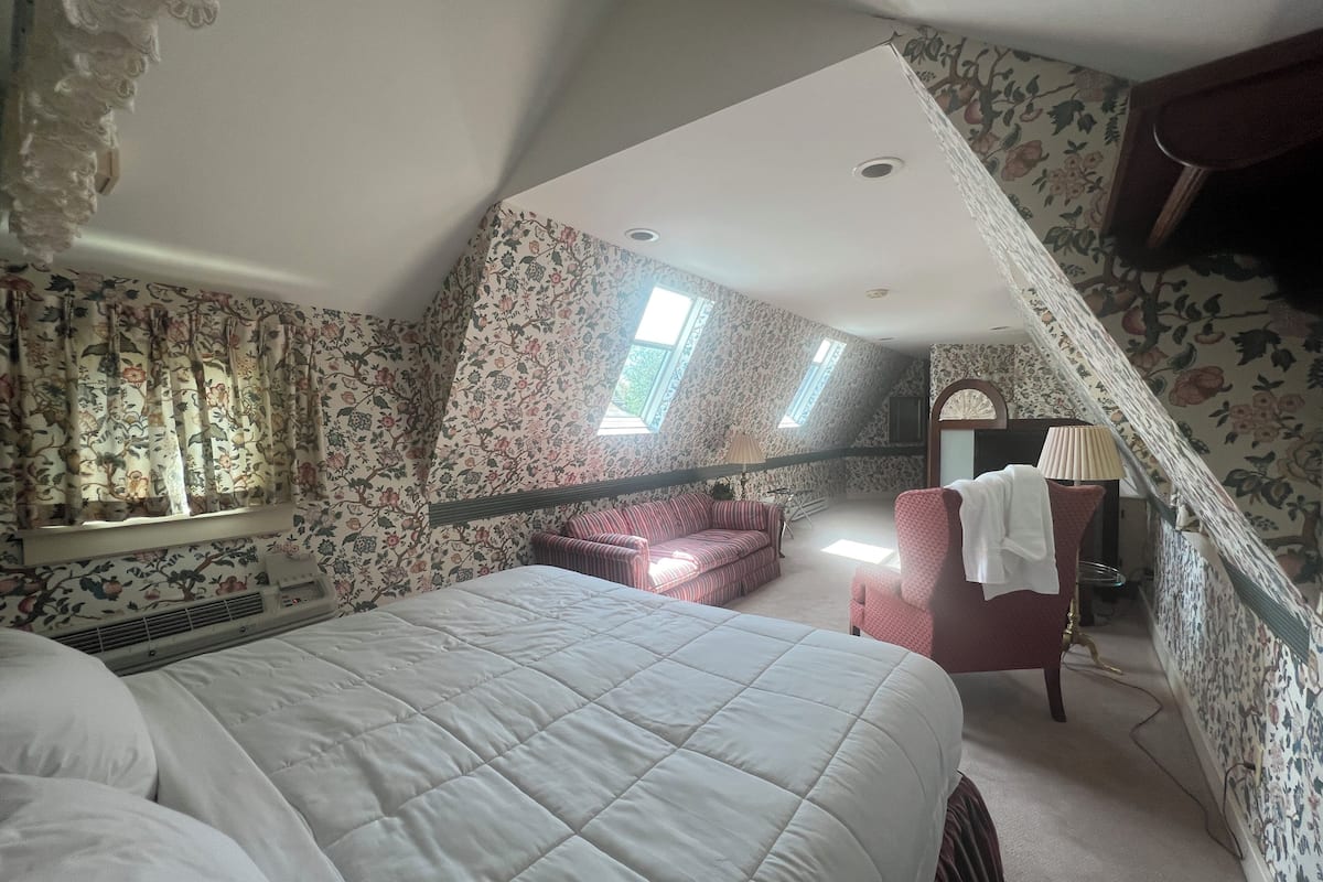 A bedroom with a large bed, a red armchair, a small table, and a window with floral wallpaper.