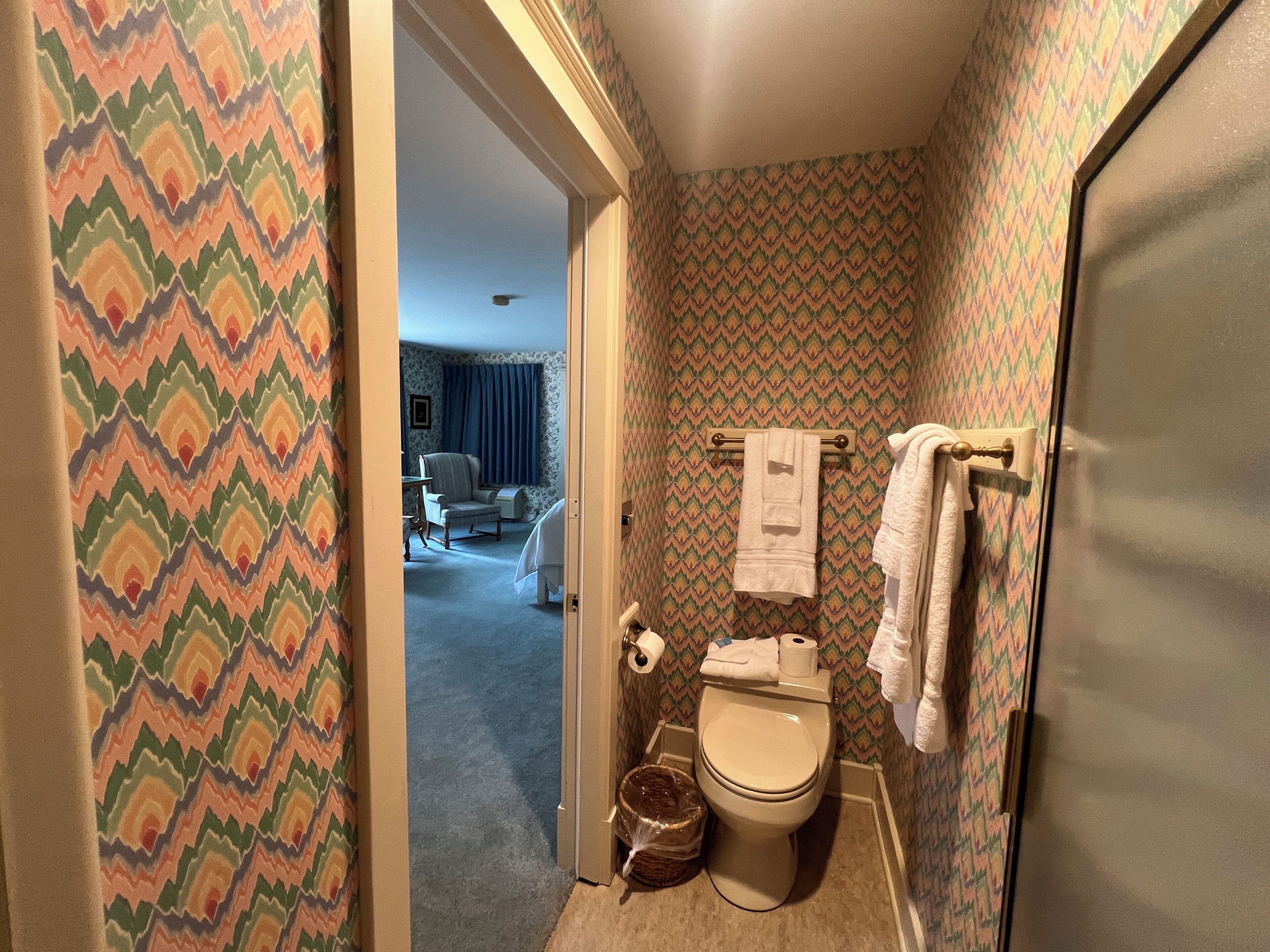 A bathroom with patterned wallpaper, a toilet, a towel rack, and a trash bin.