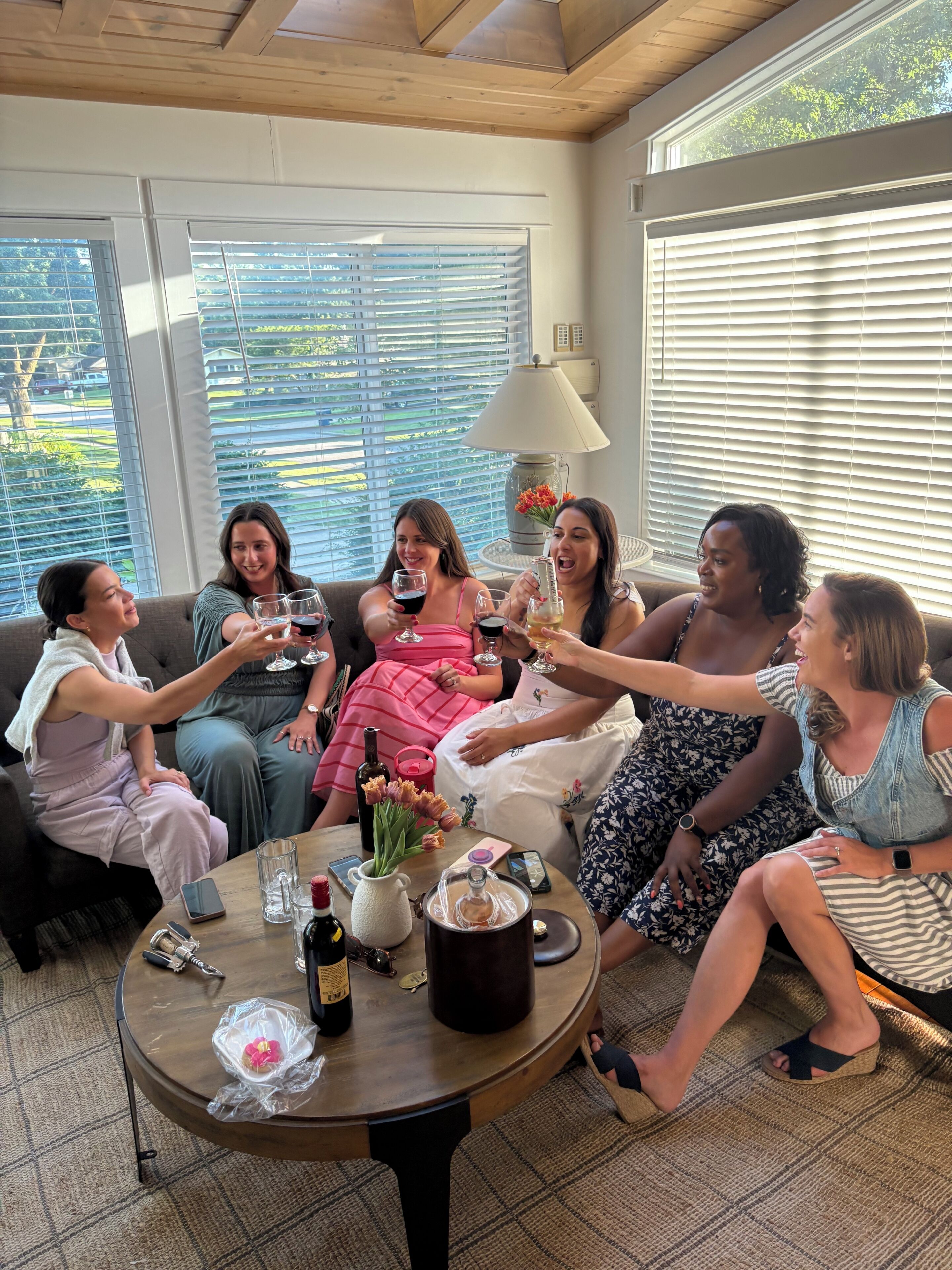 Un grupo de personas sentadas alrededor de una mesa redonda, con bebidas, en una sala con ventanas amplias y techo de vigas de madera.