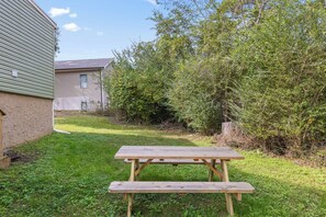 Ett picknickbord av trä i en grönskande trädgård, med ett hus och tät vegetation i bakgrunden.