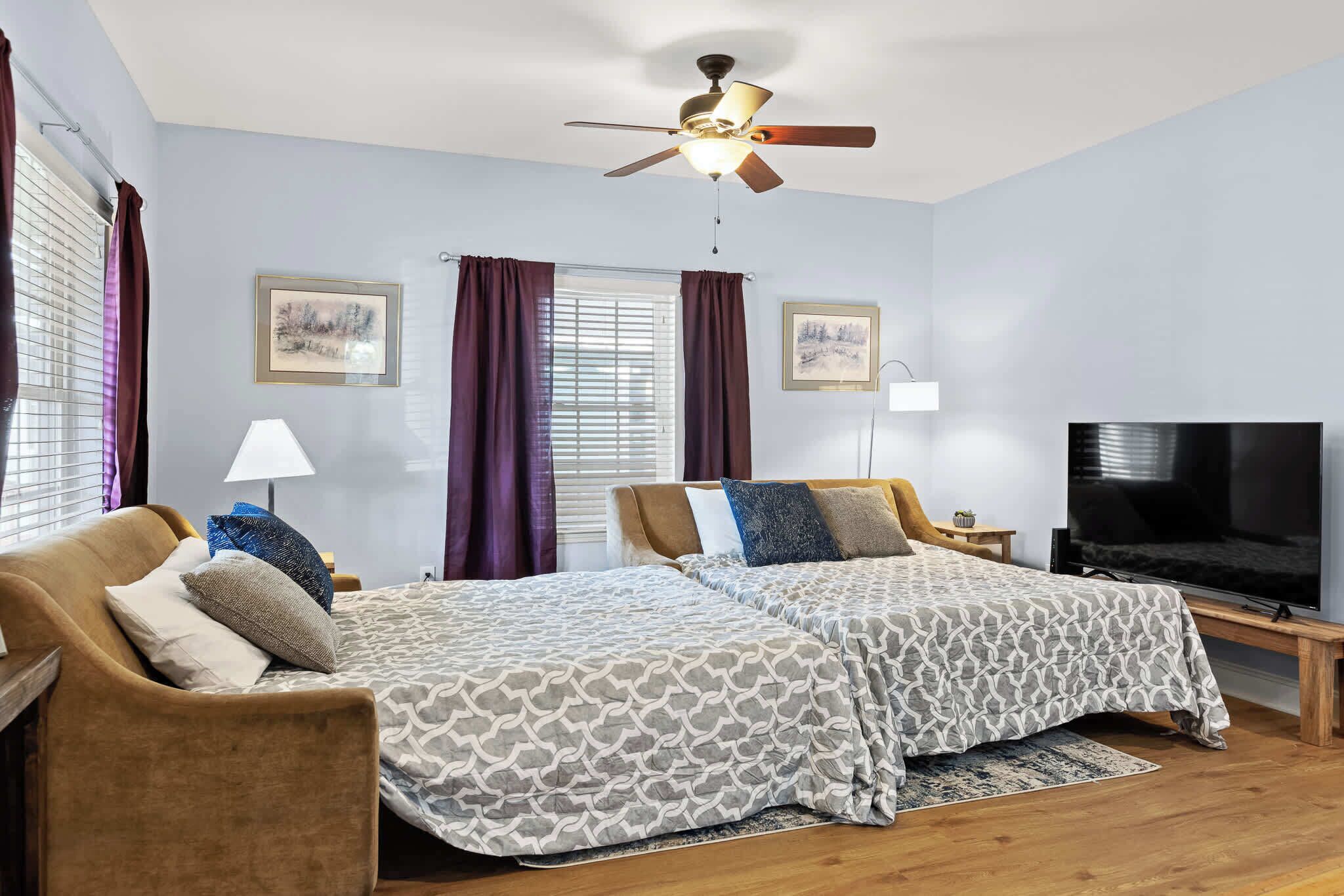 A bedroom with two beds, a ceiling fan, a TV, and artwork on the walls.