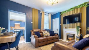 A modern living room with a leather sofa, a dining table with chairs, a fireplace, and a TV mounted on the wall.