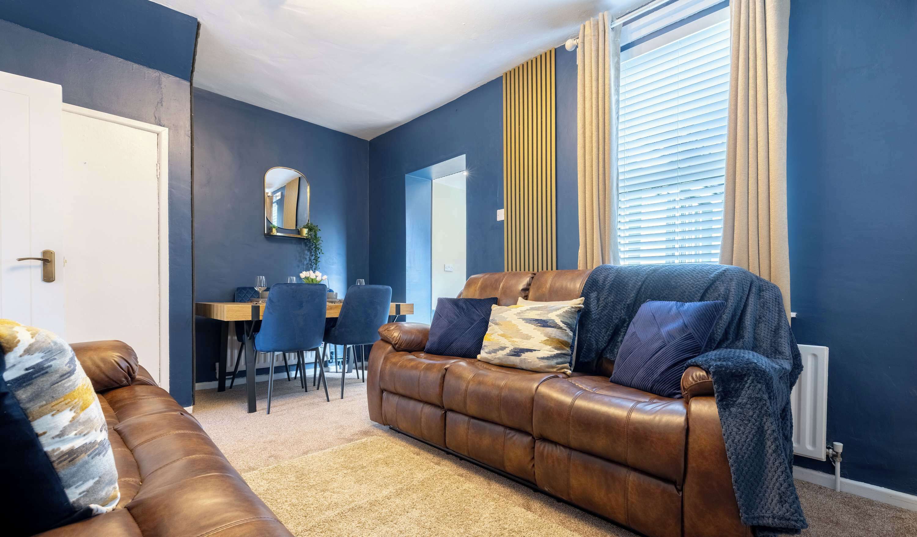 A living room with a brown leather sofa, a dining table with chairs, a mirror, and a window with blinds.