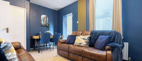 A living room with a brown leather sofa, a dining table with chairs, a mirror, and a window with blinds.