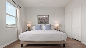 A bedroom with a bed, two bedside lamps, a framed picture on the wall, and a window with curtains.
