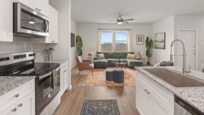 A modern kitchen with a built-in oven and microwave, a dining area with a large island and sink, and a living room with a green sofa and a view of the outdoors.