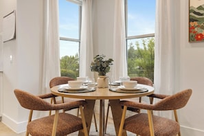 A round dining table with four wicker chairs, a vase with greenery, and white plates and cups.