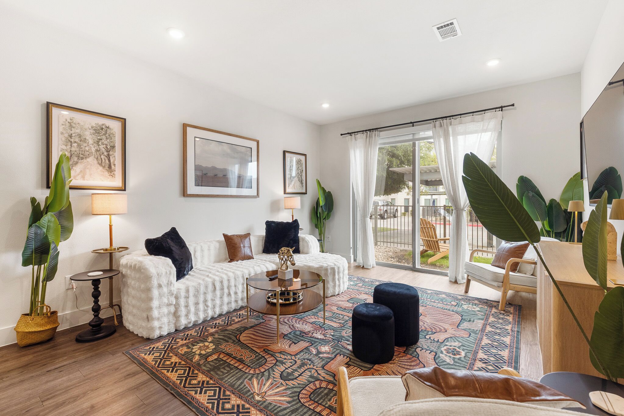Un salon moderne avec un canapé blanc, une table basse en verre et un tapis à motifs. On y trouve deux grandes plantes en pot, une lampe et un tableau encadré accroché au mur.
