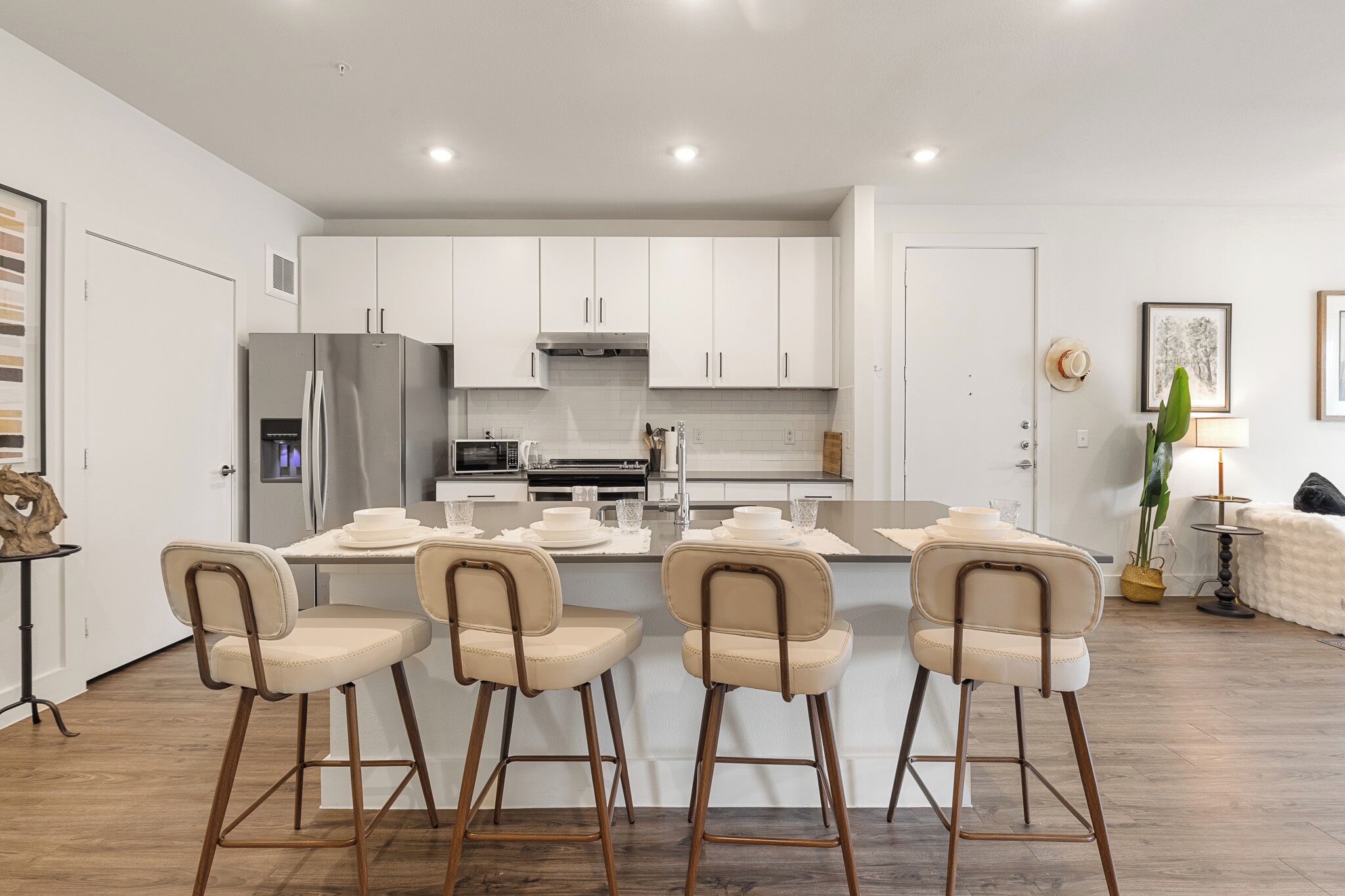 A modern kitchen with white cabinets, a central island with bar stools, stainless steel appliances, and a dining area with a table and chairs.