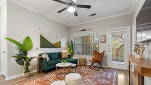Una sala de estar con un sofá verde, un ventilador de techo, una planta grande y un tapete estampado.