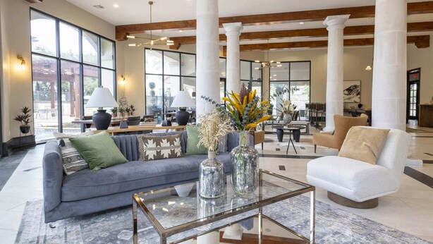 Un vestíbulo amplio con un sofá gris, un sillón blanco y una mesa de centro de vidrio. Sobre la mesa de centro hay dos jarrones con flores. La habitación tiene vigas de madera en el techo y ventanas grandes.