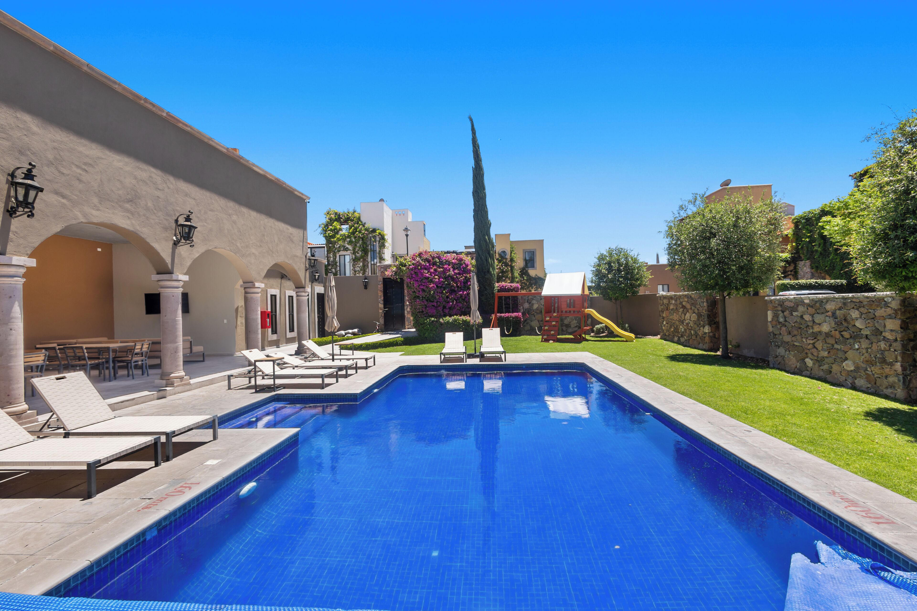 A swimming pool with lounge chairs, a stone wall, and a clear blue sky.