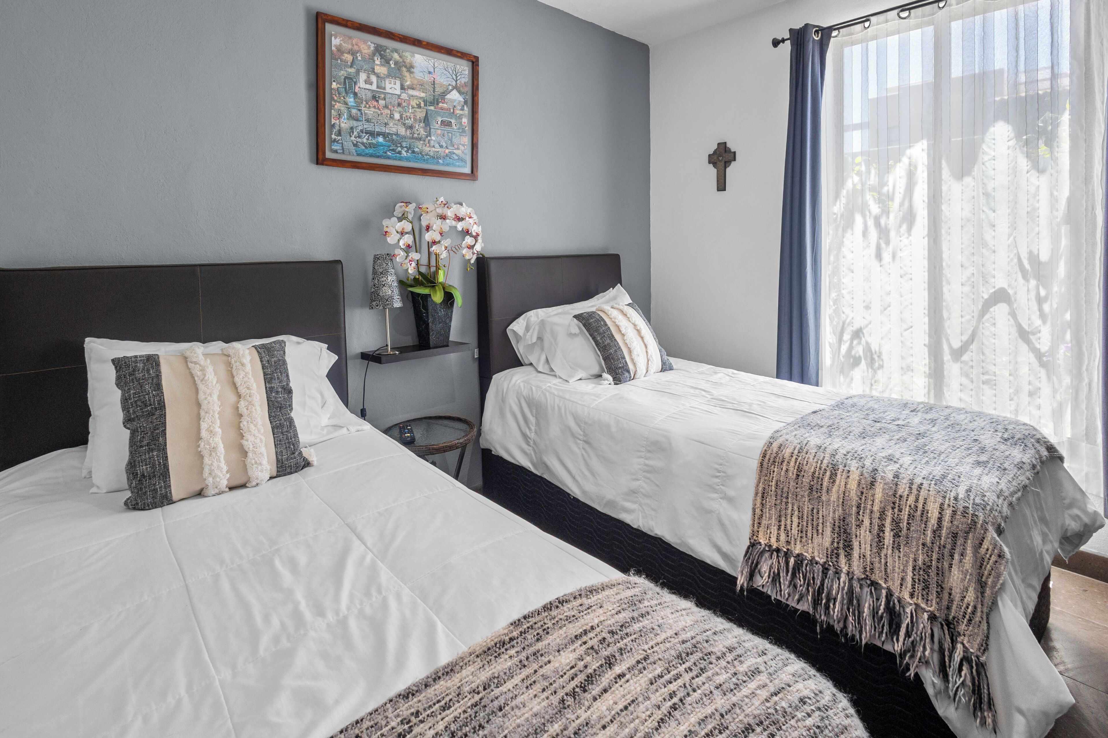 A hotel room with two beds, a nightstand with a vase of flowers, a framed picture on the wall, and a window with curtains.