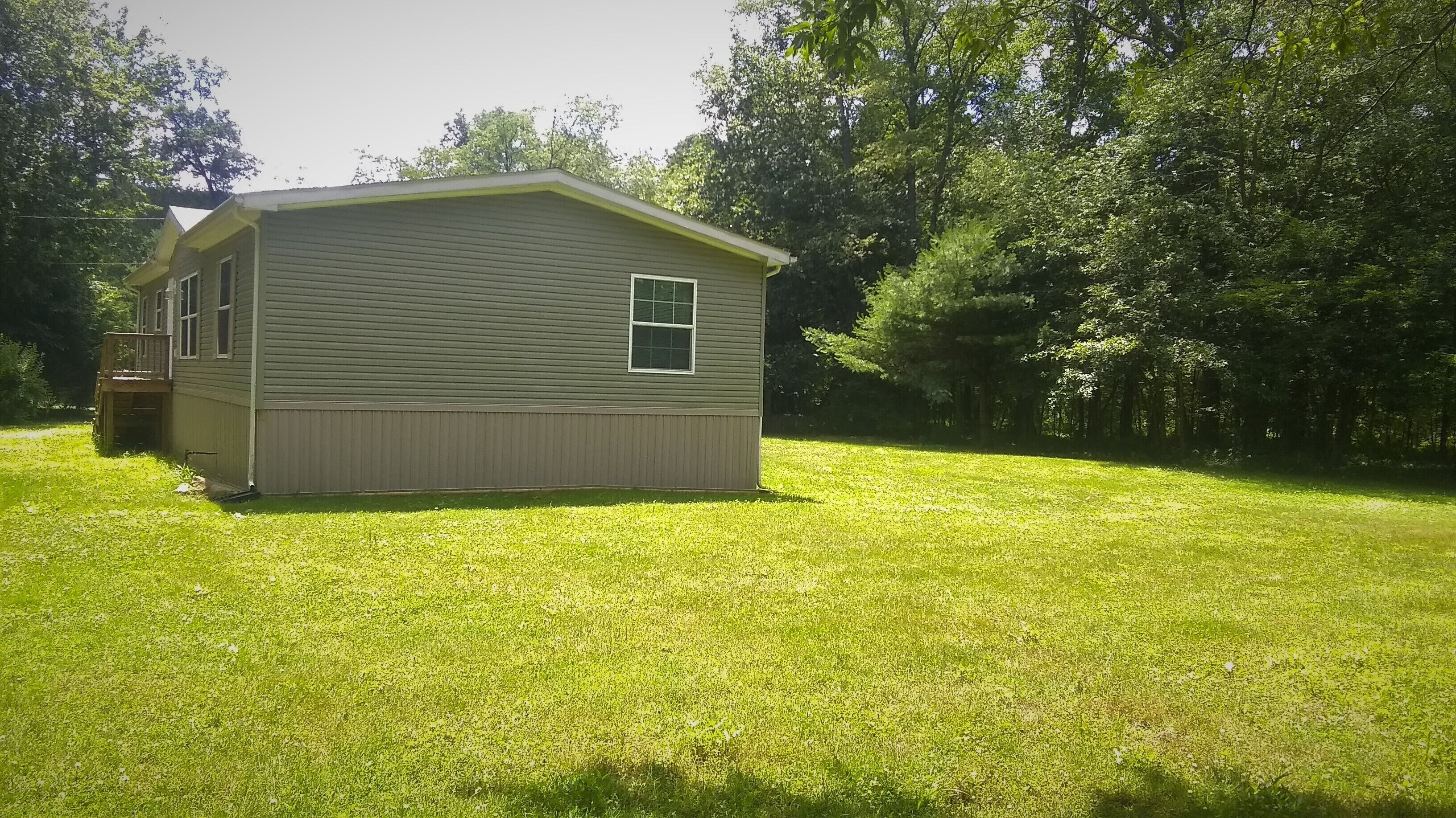 A single-story house with a front porch and a small deck, surrounded by a well-maintained lawn and dense trees.