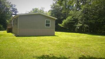 A single-story house with a front porch and a small deck, surrounded by a well-maintained lawn and dense trees.