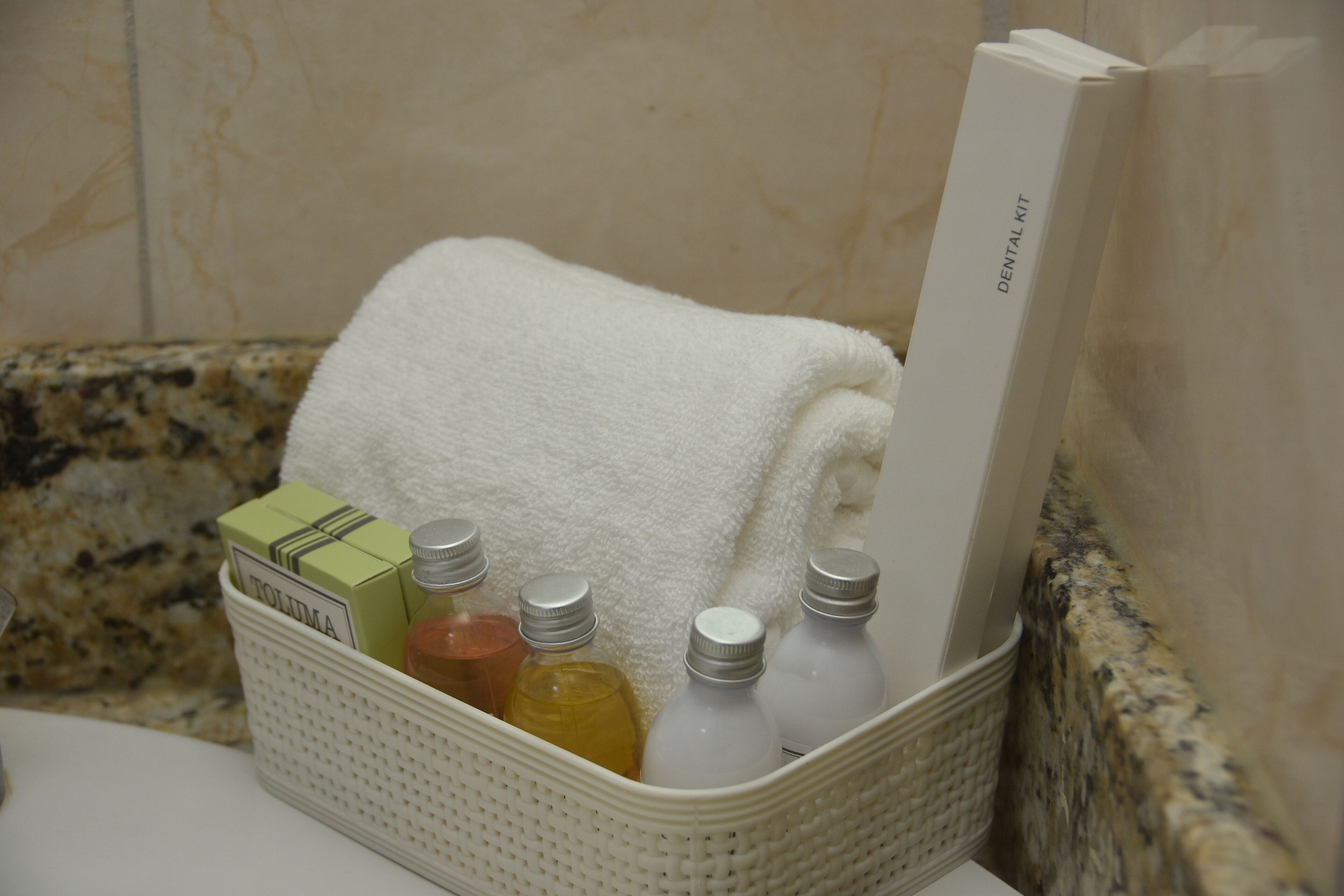 A bathroom with a white towel, a dental kit, and a basket containing toiletries.