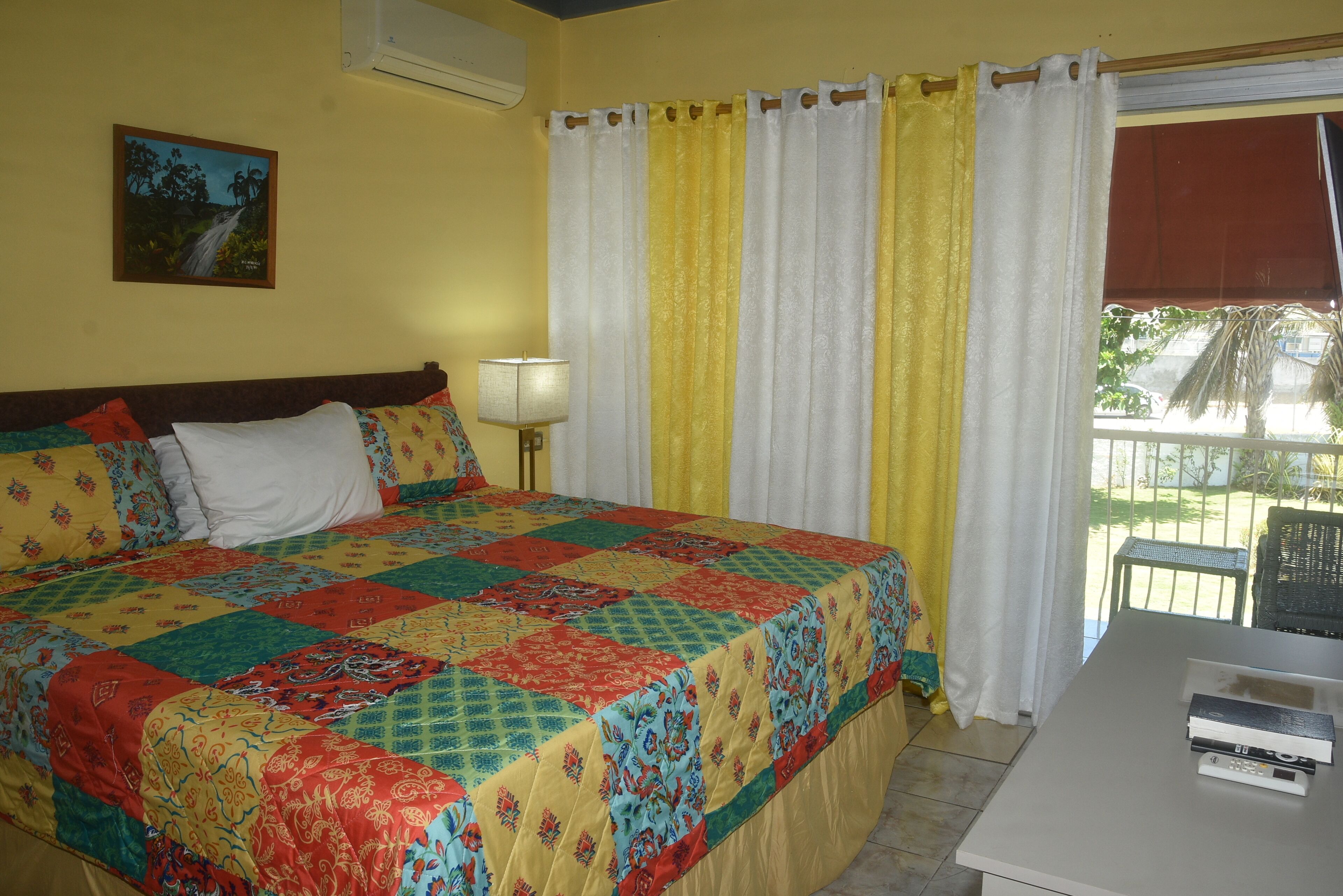 A bed with a colorful quilt, a bedside lamp, a window with curtains, and a small table with books.
