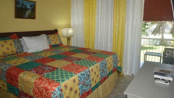 A bed with a colorful quilt, a bedside lamp, a window with curtains, and a small table with books.