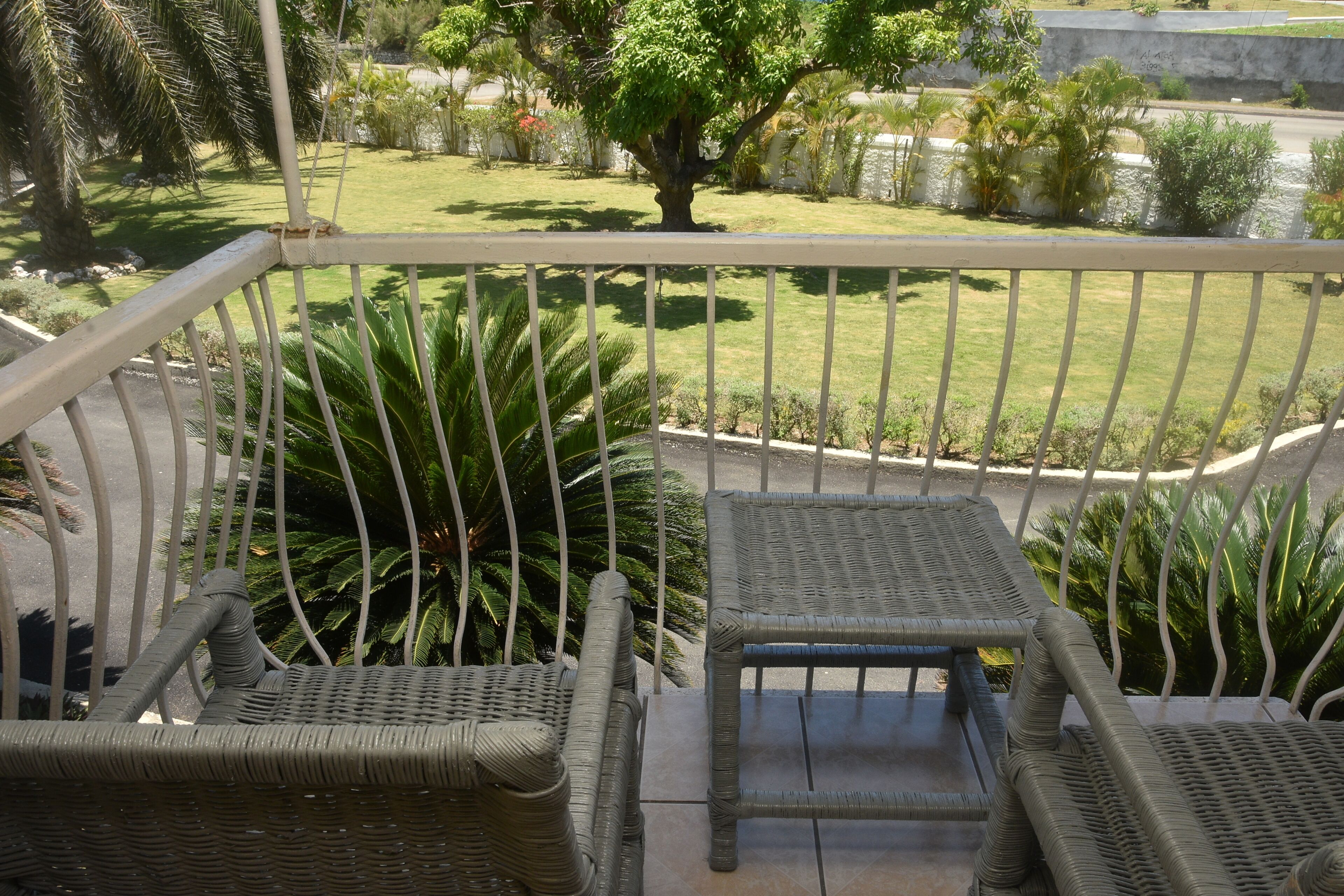 A balcony with wicker furniture, a view of a well-maintained garden, and a clear sky.