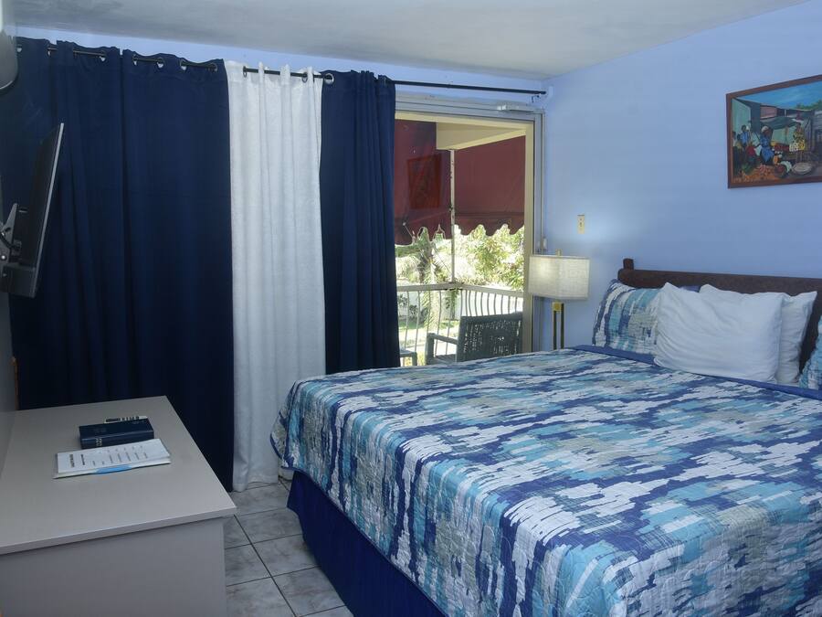 A hotel room with a bed, a desk with a lamp, a chair, a window with curtains, and a painting on the wall.