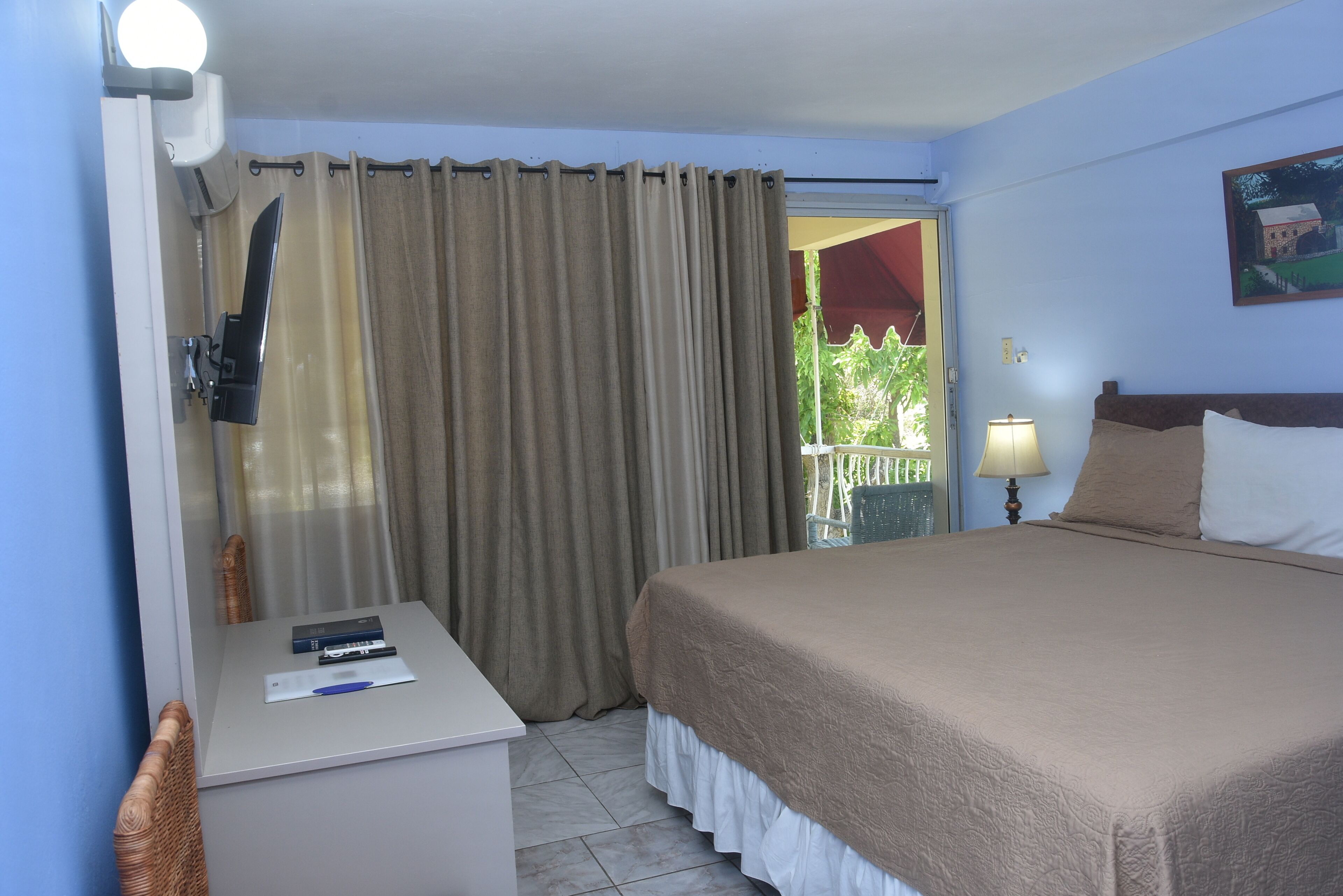 A hotel room with a bed, a desk with a lamp, a window with curtains, and a painting on the wall.