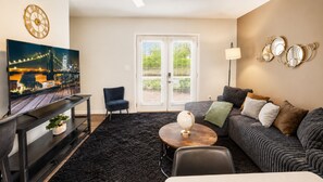 A living room with a sofa, a coffee table, a chair, a TV, a clock, and a window with blinds.