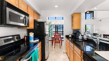 A modern kitchen with a black stove, stainless steel microwave, and refrigerator, wooden cabinets, a black countertop, and a dining area with a table and chairs.