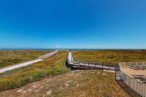 A coastal walkway with a wooden deck, a sandy area with lounge chairs, and a clear view of the ocean.