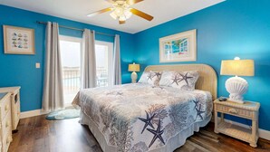 Une chambre à coucher comprenant un lit, deux tables de chevet, un ventilateur de plafond, une fenêtre avec des rideaux et une vue sur l’extérieur.