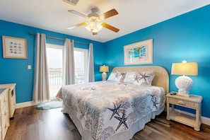 A bedroom with a bed, two bedside tables, a ceiling fan, a window with curtains, and a view of the outdoors.
