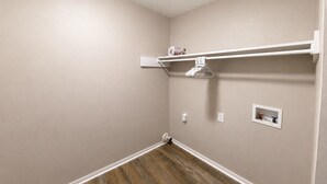 A small, empty room with beige walls, a wooden floor, a white shelf with a towel, and two electrical outlets. - Olive & Elm Hoover | Cozy Modern Escape *1Bed* (Hoover)