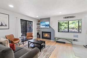 Ein Wohnzimmer mit Kamin, einer Couch, zwei Sesseln, einem Couchtisch und einem großen Fenster mit Blick ins Grüne.