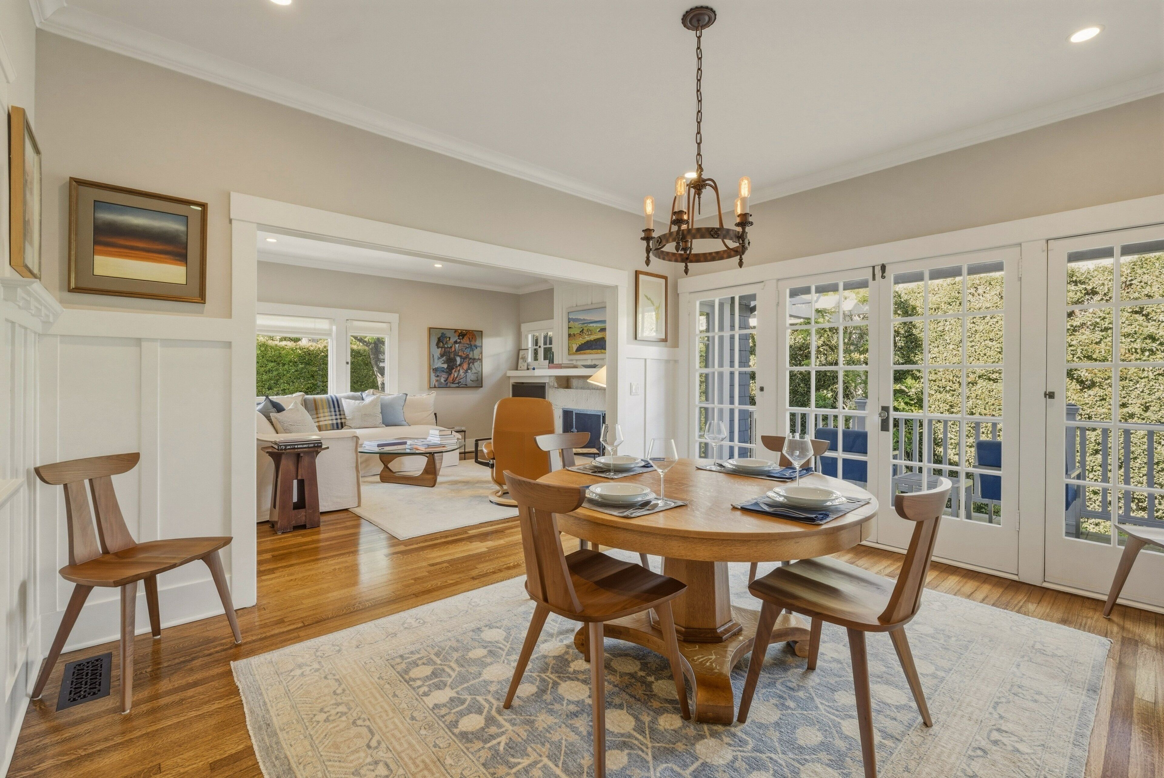 A dining area with a round table, chairs, and a chandelier. There is a sofa and a coffee table in the background.